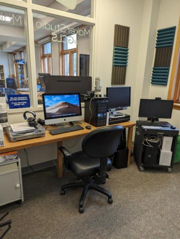 interior of the library's digital media lab. On a computer desk is a Mac with podcast recording equipment, a VHS to DVD transfer device, and a gaming computer is on a a cart in the corner.